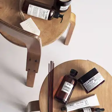 Två runda träpallar med hudvårdsprodukter från L:A BRUKET placerade på dem. Produkterna inkluderar bruntonade flaskor, en vit tvål med snöre, en burk med kroppskräm och en tub med handkräm. Doftpinnar ligger bredvid en flaska med etiketten "No 202 CORIANDER". Bilden har en minimalistisk och stilren komposition med varma trätoner och en ljus bakgrund.