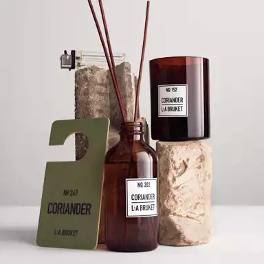 En stilren produktuppställning med L:A BRUKET-produkter i doften Coriander. På bilden syns en brun glasflaska med doftpinnar (No 202), ett mörkbrunt doftljus (No 152), och en grön dörrskylt med texten No 247 CORIANDER. Produkterna är arrangerade tillsammans med råa betong- och stenblock, samt en genomskinlig tändare. Bakgrunden är ljus och minimalistisk.