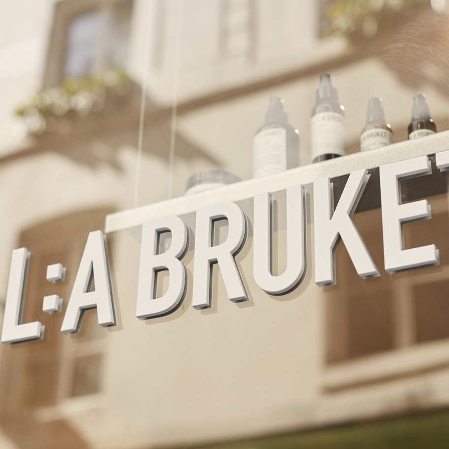 Närbild av en skylt med texten "L:A BRUKET", monterad på ett glasfönster. Skylten är i vita, upphöjda bokstäver. I bakgrunden syns reflektioner av en byggnad med fönster och ljusa fasader, samt några hudvårdsprodukter på en hylla innanför glaset. Ljuset är varmt och naturligt.
