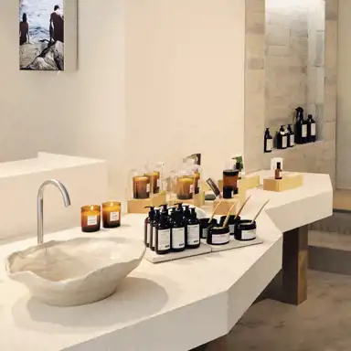 En butiksmiljö med en minimalistisk design där L:A BRUKET-produkter är arrangerade på ett vitt, rustikt bord med träben. På bordet står ett handfat i organisk form, en modern kran, bruntonade flaskor, doftljus och burkar med hudvårdsprodukter. I bakgrunden syns en vägg med inbyggda hyllor där fler flaskor och produkter är placerade, samt en tavla med en bild på två personer vid vatten. Ljuset är varmt och skapar en inbjudande atmosfär.