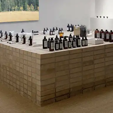 En butiksmiljö med en stor rektangulär display byggd av beige tegelstenar, där L:A BRUKET-produkter är prydligt arrangerade. Flaskor, burkar och askar med hudvårdsprodukter står i grupper över hela ytan. I bakgrunden syns en tavla med ett landskapsmotiv av ett fält och en skog. Atmosfären är minimalistisk och naturlig.