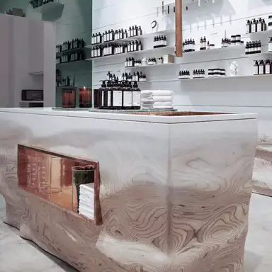 En stilren butiksmiljö med en central disk i ljust trä med synliga ådringar och en inbyggd kopparhylla som innehåller glasbehållare och vikta handdukar. På disken står bruntonade flaskor med L:A BRUKET-produkter, vita handdukar och tända ljus. Bakom disken finns vita hyllor fyllda med flaskor och burkar i en minimalistisk uppställning. Ljussättningen skapar en ren och modern atmosfär.