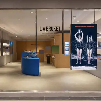 Butiksfasaden till L:A BRUKET med stora glasfönster och en modern, minimalistisk inredning. En blå rund disk med ett handfat står i centrum, omgiven av produkter och träpaneler. På fönstret syns texten "Natural and organic skincare made on the west coast of Sweden." Till höger hänger en stor digital skärm som visar en bild av två personer i baddräkter som häller iskallt vatten över sig, med texten "Winter rituals of well-being. Inspired by nature." Ljuset inne i butiken är varmt och inbjudande.