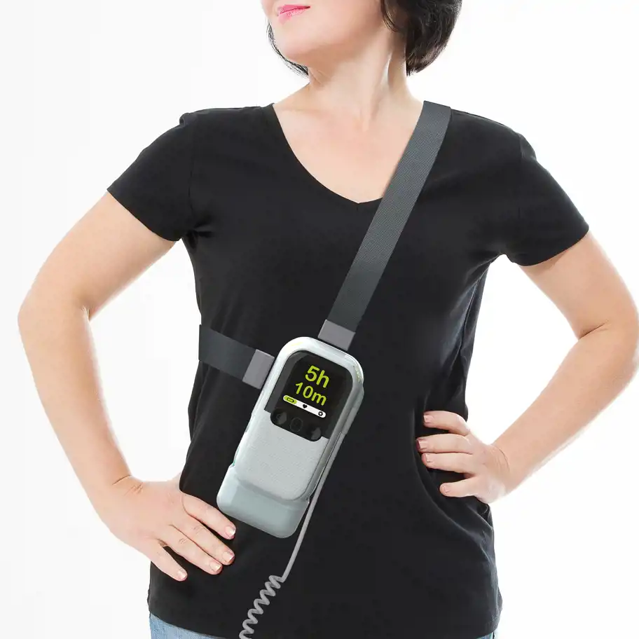En person i svart t-shirt bär en medicinsk enhet över axeln och runt midjan med hjälp av en justerbar rem. Enheten har en digital skärm som visar "5h 10m" tillsammans med olika indikatorer. En spiralformad sladd är ansluten till enhetens nederdel.