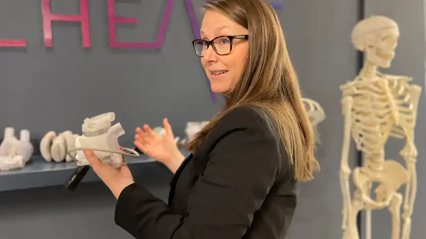 En kvinna med långt brunt hår och glasögon bär en svart kavaj och håller upp en medicinsk prototyp av ett artificiellt hjärta medan hon förklarar något. Hon står framför en mörkblå vägg med företagets logotyp i lila och rosa nyanser. I bakgrunden syns fler hjärtmodeller på en hylla samt ett skelett.