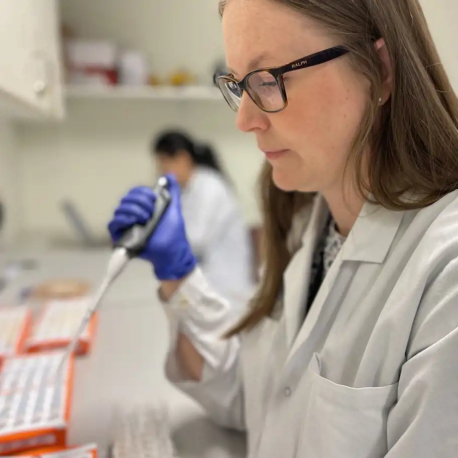 En kvinna i vit laboratorierock och glasögon pipetterar en vätska i ett laboratorium. Hon bär blå skyddshandskar och ser koncentrerad ut. I bakgrunden arbetar en annan forskare, också i vit rock, vid ett labbord med provrör och utrustning.