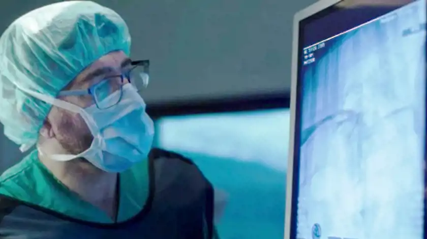 En kirurg iförd operationsmössa, munskydd och skyddsglasögon studerar en röntgenbild på en monitor i en operationssal. Bilden på skärmen visar en anatomisk struktur i blå toner. Bakgrunden är suddig, men en annan skärm och medicinsk utrustning syns.