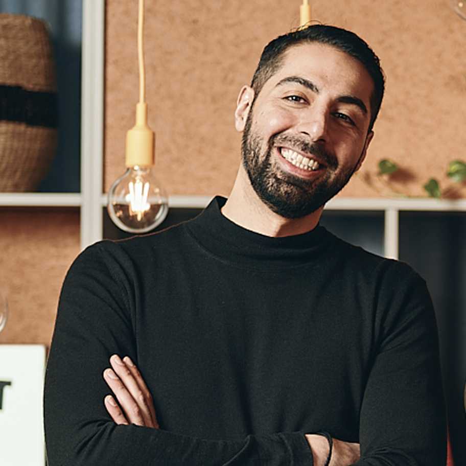 En leende man med kort mörkt hår och skägg bär en svart polotröja och står med armarna korsade framför sig. Bakgrunden har en varm, modern inredning med en glödlampa som hänger från taket, växter och hyllor med dekorativa föremål.