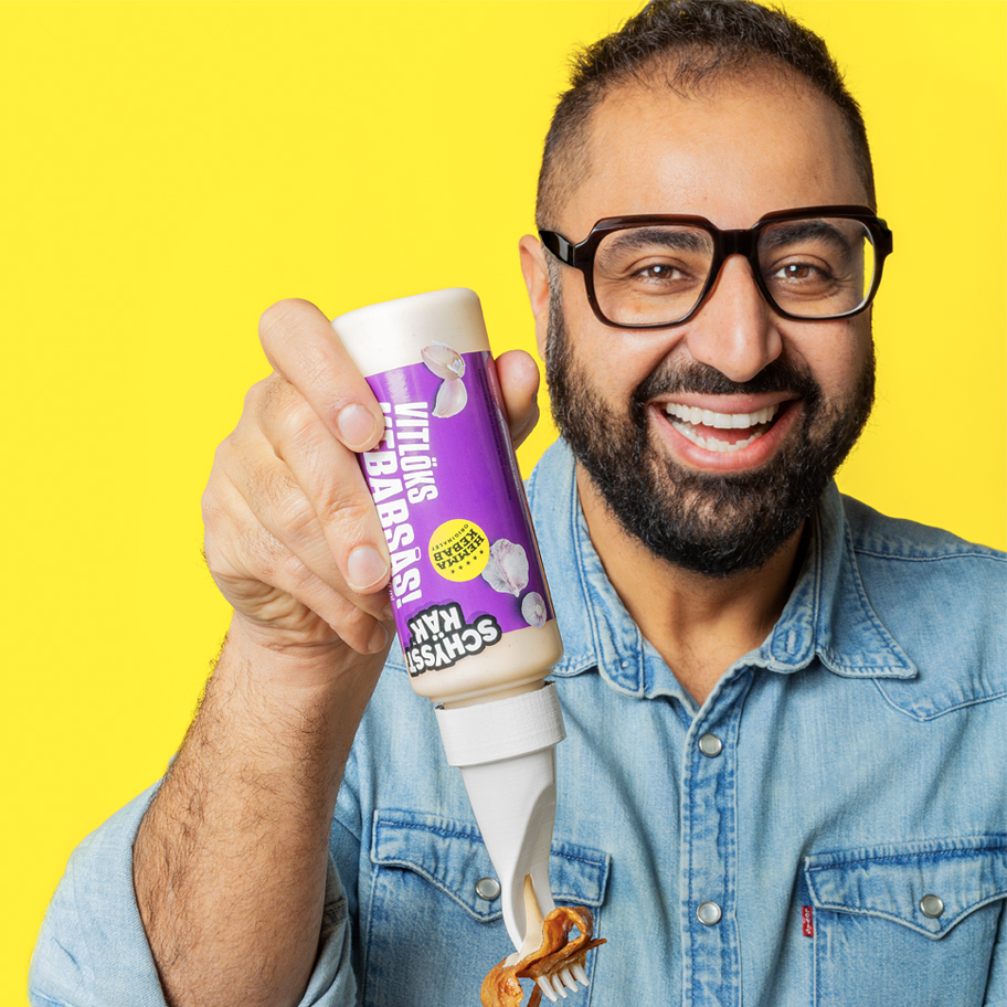 En leende man med kort mörkt hår, skägg och stora glasögon bär en jeansskjorta och håller i en flaska vitlökssås. Han trycker ut såsen på en bit mat med hjälp av en innovativ flasköppning som fungerar som en sked. Bakgrunden är starkt gul, vilket ger bilden en färgstark och lekfull känsla.