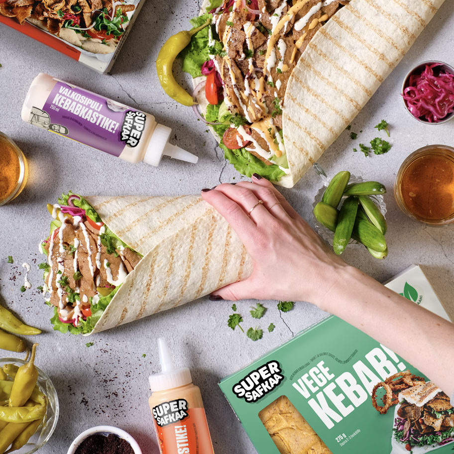 En matbild med två stora rullar av vegansk kebab i tunnbröd, fyllda med sallad, tomat, rödlök och såser. En hand med röda naglar och guldringar håller i en av rullarna. Runt omkring ligger ingredienser som feferoni, inlagd rödkål, små gurkor och såsflaskor med texten "Super Safkaa". På bilden syns också en förpackning med texten "Vege Kebab!", samt ett glas med en orange dryck. Bakgrunden är en ljusgrå stenbänk med färska korianderblad strödda över.