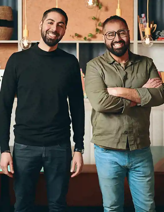 Två leende män står i ett modernt inrett kontor eller restaurangmiljö. Den ena mannen bär en svart polotröja och jeans, medan den andra bär en grön skjorta och blå jeans. De står framför en hylla fylld med böcker, växter och dekorativa detaljer. Bakom dem hänger flera glödlampor med gula sladdar. På en hylla syns en ljusskylt med texten "SCHYSST KÄK", och det finns böcker med titlar som "Pizza" och "India". Miljön har en modern och inbjudande känsla.