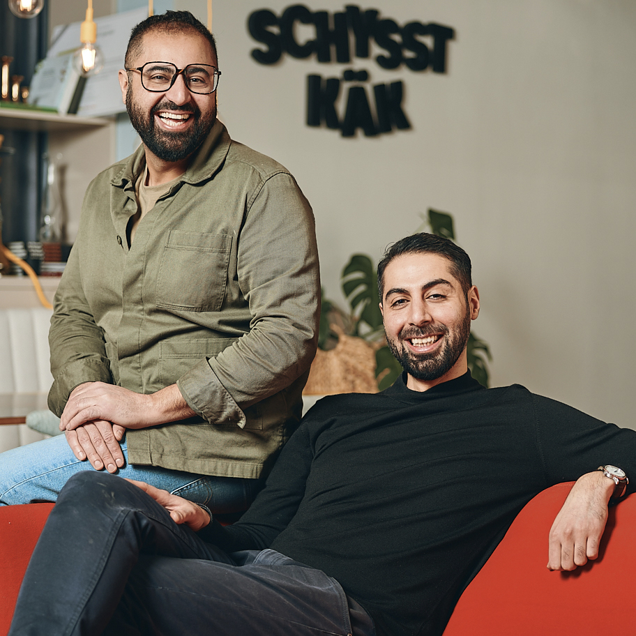 Två leende män sitter avslappnat i en modern inredd miljö med en skylt i bakgrunden som säger "Schysst Käk". Mannen till vänster bär glasögon och en grön skjorta, och lutar sig lätt framåt med händerna vilande på sina knän. Mannen till höger ligger avslappnat i en röd fåtölj, klädd i en svart tröja och mörka byxor, och ler stort mot kameran. Bakgrunden innehåller växter och hyllor med böcker och dekorativa element.