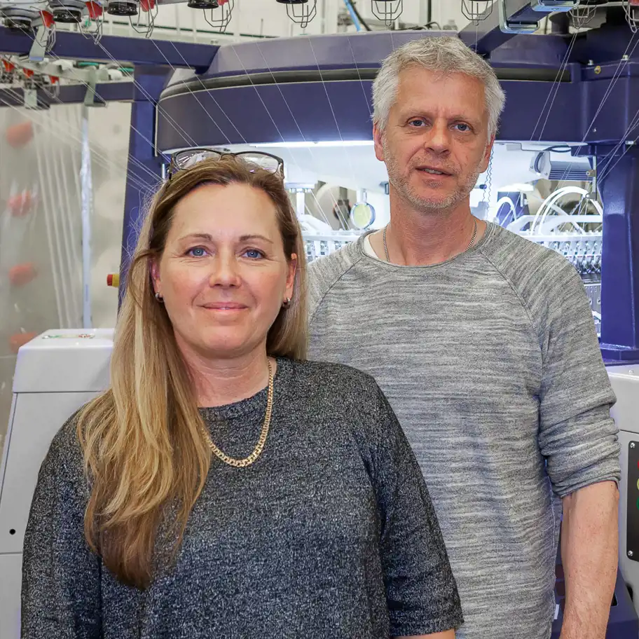 Detta är en bild av två personer i en industriell miljö, sannolikt i en textil- eller tillverkningsfabrik. De står framför en avancerad maskin som verkar vara en typ av stickmaskin eller liknande utrustning för textilproduktion. Båda personerna är klädda i avslappnade, men professionella kläder och ser ut att vara involverade i produktionen eller företaget bakom verksamheten. Maskinen bakom dem är fylld med trådar och mekaniska komponenter, vilket indikerar en högteknologisk och automatiserad process.