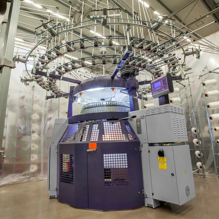 En stor cirkulär stickmaskin i en industriell miljö. Maskinen har en robust metallram och ett system av garnspolar och matarledningar ovanpå, som fördelar garn till maskinen. Maskinen är försedd med ett kontrollpanel på sidan och är omgiven av hyllor med garnspolar i bakgrunden. Ljuset i rummet är jämnt och ger en ren och modern känsla.