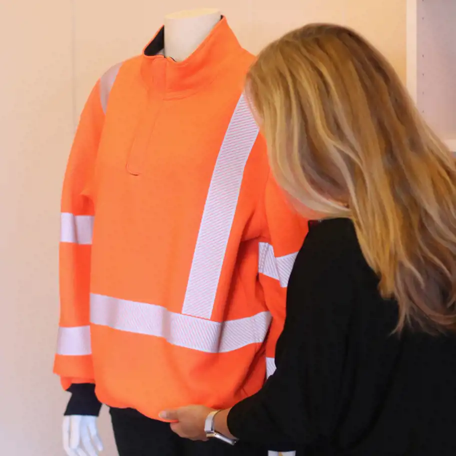 Den här bilden visar en person som granskar en högsynlighets-tröja på en skyltdocka. Tröjan är fluorescerande orange med reflekterande ränder, vilket är typiskt för arbetskläder inom bygg- och transportsektorn där synlighet och säkerhet är avgörande.  Personen på bilden verkar inspektera eller analysera materialet och designen, vilket kan tyda på att de arbetar inom textil- eller säkerhetsklädesindustrin. Det kan vara en del av en produktutvecklingsprocess där kvalitet, funktionalitet och säkerhetsstandarder bedöms.