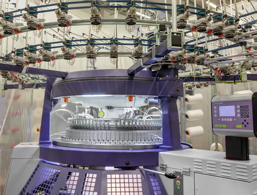 Närbild av en cirkulär stickmaskin i en industriell miljö. Maskinens övre sektion visar ett komplext system av garnmatare och rullar, där garnen leds ner mot stickningsområdet. Den centrala delen har en metallisk rund stickenhet med nålar och tillbehör, medan en elektronisk kontrollpanel med en digital skärm syns på sidan. Garnspolar är synliga i bakgrunden, monterade på hyllor. Ljusa och rena omgivningar förstärker det tekniska intrycket.