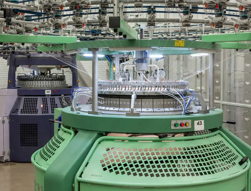 Två cirkulära stickmaskiner i en modern industrimiljö. Den närmaste maskinen är grön med en rund stickningsenhet och garnmatningssystem ovanför. Garn träs genom flera ledningar mot maskinens centrum, där stickningen sker. I bakgrunden syns en lila maskin med liknande konstruktion. Ljuset i rummet är starkt och jämnt, vilket skapar en ren och tekniskt avancerad atmosfär.