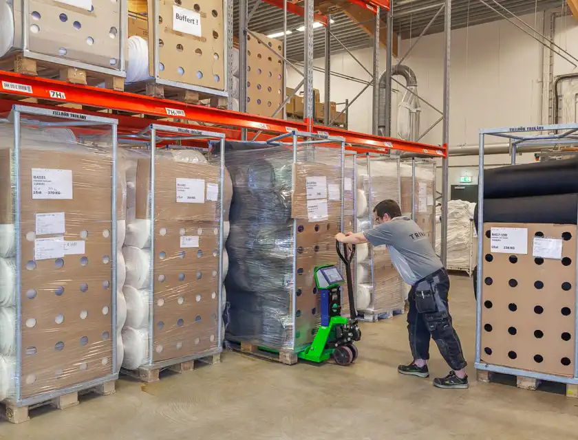 Bilden visar en lagerlokal för textilproduktion, där stora rullar av garn och textilmaterial förvaras på pallställ och metallramar. En anställd använder en elektrisk pallyftare med en digital skärm för att hantera materialet effektivt.  Etiketterna på förpackningarna tyder på att detta är ett organiserat lager där olika typer av textilprodukter hanteras och spåras noggrant. De perforerade kartongförpackningarna runt rullarna kan indikera att materialen är skyddade samtidigt som de är lättillgängliga för produktion.  Den moderna lagerhanteringen och arbetsklädseln hos den anställde antyder en professionell och välorganiserad verksamhet, troligen kopplad till en större textilproduktionskedja.