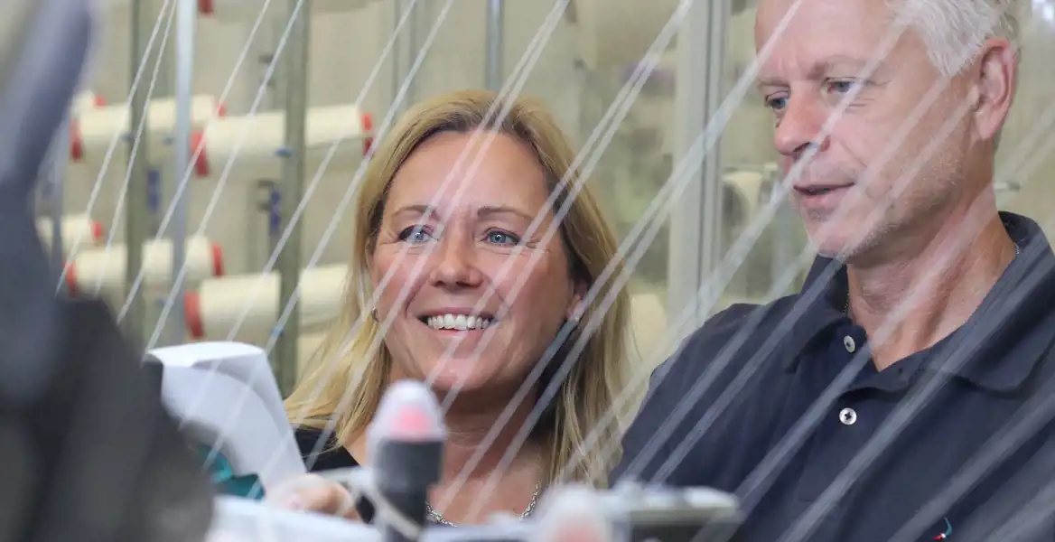 Bilden visar två personer i en textilproduktionsmiljö, där trådar sträcker sig i förgrunden. De verkar observera eller inspektera processen, vilket kan tyda på att de är involverade i tillverkning eller kvalitetskontroll av textilier.  Den glada och engagerade uttrycket hos kvinnan och mannens fokuserade blick antyder en passion för arbetet, kanske som en del av en innovativ textilindustri. Bakgrunden med trådar och maskiner bekräftar att detta är en avancerad tillverkningsmiljö, troligen relaterad till garn- eller tygproduktion.