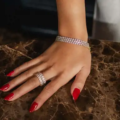 En hand med rödmålade naglar vilar på en mörk marmoryta. Handen är smyckad med ett glittrande armband och en bred ring med diamanter, båda i en elegant design som fångar ljuset och skapar en lyxig känsla.