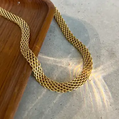 Ett guldhalsband med en flätad design ligger delvis på en trälåda och sträcker sig ut över en betongyta. Solljuset reflekteras genom kedjan och skapar ett vackert spektrum av ljus och skuggor på underlaget.