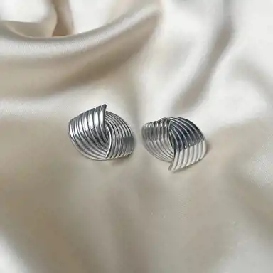 Ett par silverörhängen med en elegant, böljande design ligger på ett krämfärgat sidenliknande tyg. Deras blanka yta reflekterar ljuset och framhäver de mjuka formerna och det sofistikerade uttrycket.