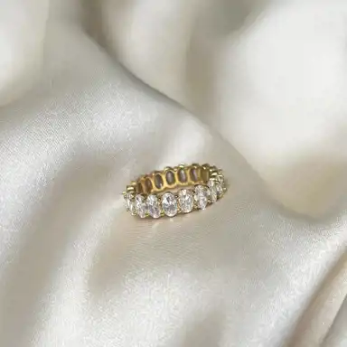 En elegant guldring med en rad gnistrande diamanter ligger på ett krämfärgat sidenliknande tyg. Ljuset fångas i stenarna och skapar en lyxig och sofistikerad känsla.