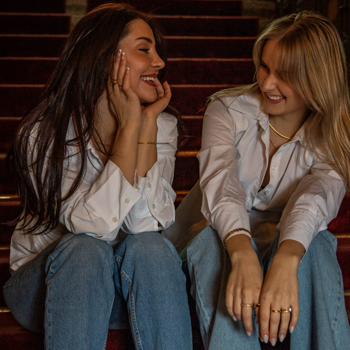 Två unga kvinnor, Albina Elshani och Alexandra Öhrvall, grundarna av Alora Avenues, sitter på en röd sammetsklädd trappa, klädda i vita skjortor och jeans. De ler och tittar på varandra i en varm och avslappnad stund. Båda bär eleganta guldsmycken, inklusive armband, ringar och halsband, som kompletterar deras stil.