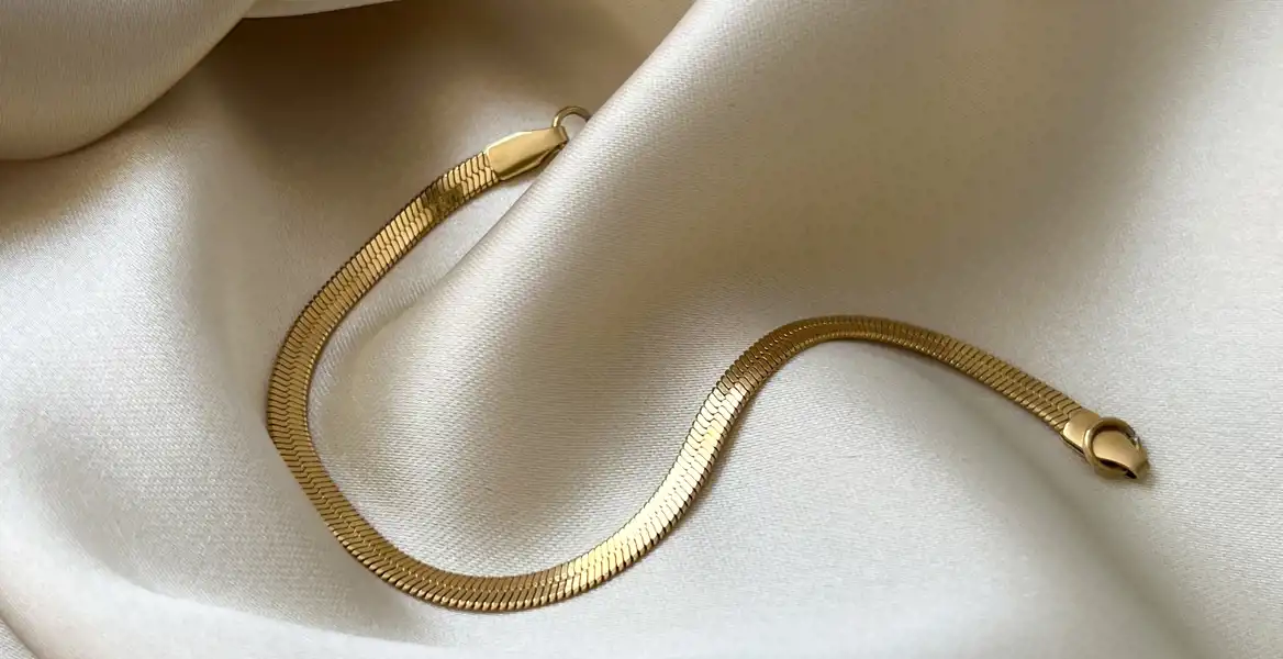 Ett guldarmband i en elegant herringbone-design ligger på ett krämfärgat sidenliknande tyg. Ljuset reflekteras mjukt på armbandet, vilket framhäver dess glänsande yta och detaljerade struktur.
