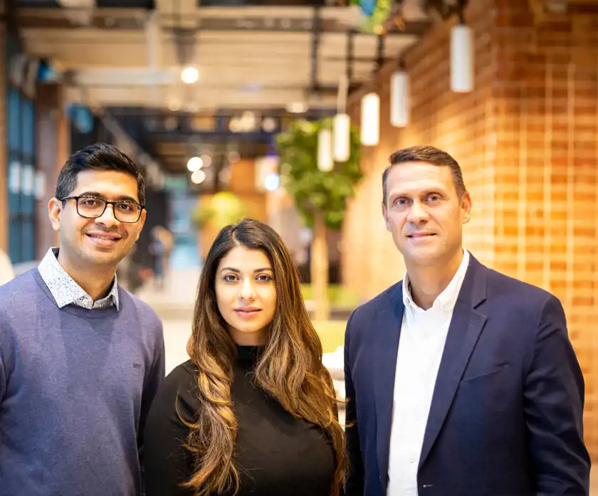 Den här bilden visar en grupp på tre professionella individer i en modern, ljus kontorsmiljö med en stilren och inbjudande atmosfär. Personerna är klädda i affärsmässiga men avslappnade kläder, vilket förmedlar en balans mellan professionalitet och tillgänglighet.  Personen till vänster bär glasögon och en stilren blå tröja med en vit skjorta under, vilket ger ett smart-casual intryck. Personen i mitten har långt, vågigt hår och bär en elegant svart topp, vilket ger en sofistikerad och självsäker framtoning. Personen till höger är klädd i en mörk blazer och vit skjorta, vilket signalerar en mer formell affärsstil. Bakgrunden med gröna växter och modern inredning skapar en varm och dynamisk känsla, och gruppens kroppsspråk förmedlar en känsla av sammanhållning och professionalism.
