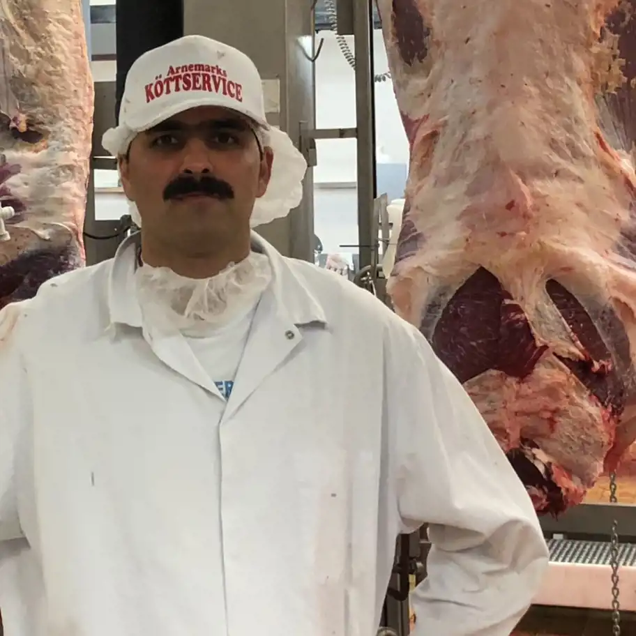 En man i en vit slaktrock och en keps med texten "Arnemarks Köttservice", vilket indikerar att han arbetar inom köttindustrin. Han har en mustasch och bär en hårnät och skäggnät för hygien. Bakom honom hänger stora köttstycken i en slakterimiljö. Hans uttryck är seriöst och professionellt, och miljön ser steril och välorganiserad ut, typisk för en modern köttbearbetningsanläggning.