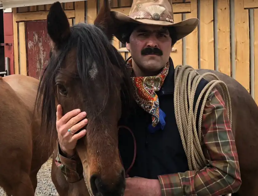 En man i cowboy-inspirerad klädsel tillsammans med en häst. Han bär en bredbrättad hatt med dekorationer, en färgglad bandana runt halsen och en rutmönstrad skjorta. Över axeln har han en lasso, vilket förstärker den westerninspirerade stilen. Mannen har en allvarlig och fokuserad blick, och han håller hästens huvud med en hand, vilket skapar en känsla av närhet och samspel mellan dem. Bakgrunden består av en trävärld som påminner om en ranch eller ett stall.