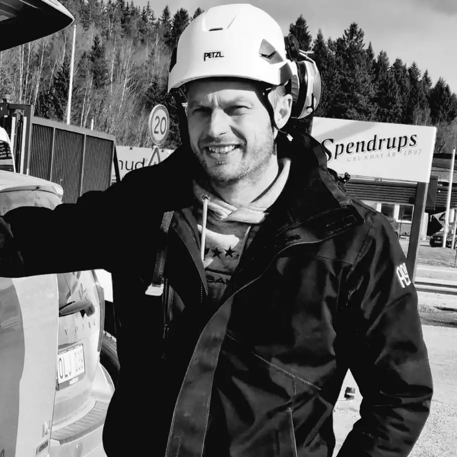 Svartvit bild på en man som bär hjälm och arbetsjacka med logotypen 'ASiL' på ärmen. Han står vid en bil med ena armen lutad mot bakluckan och ler mot kameran.