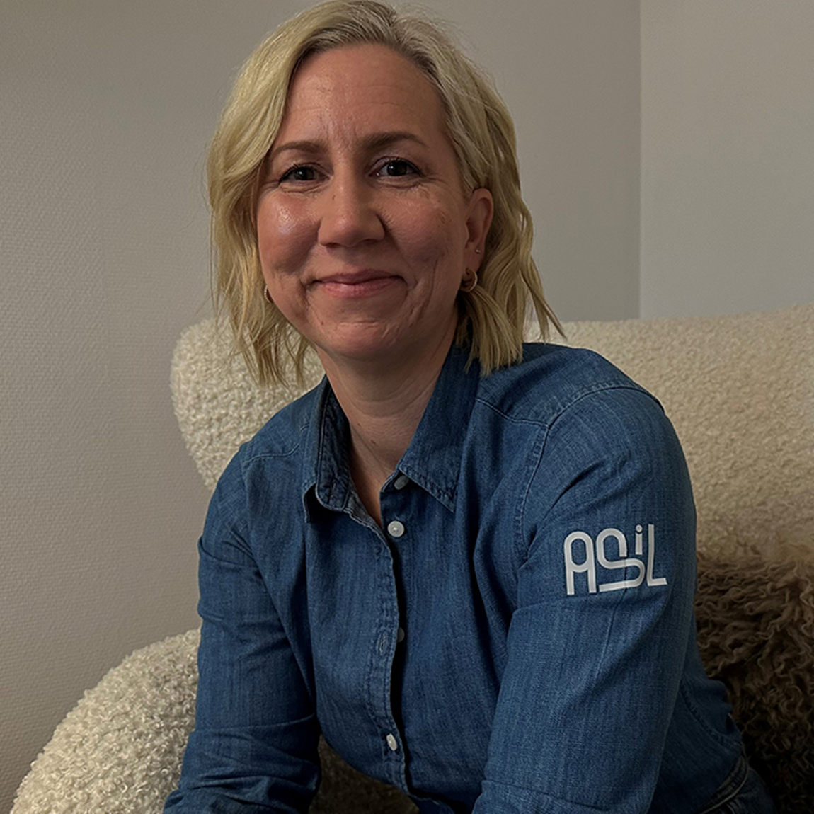 Porträtt av en kvinna med blont, axellångt hår som ler mot kameran. Hon sitter i en ljus fåtölj och bär en jeansskjorta med ASL-logotypen på ärmen. Bakgrunden är enkel med ljusa väggar.
