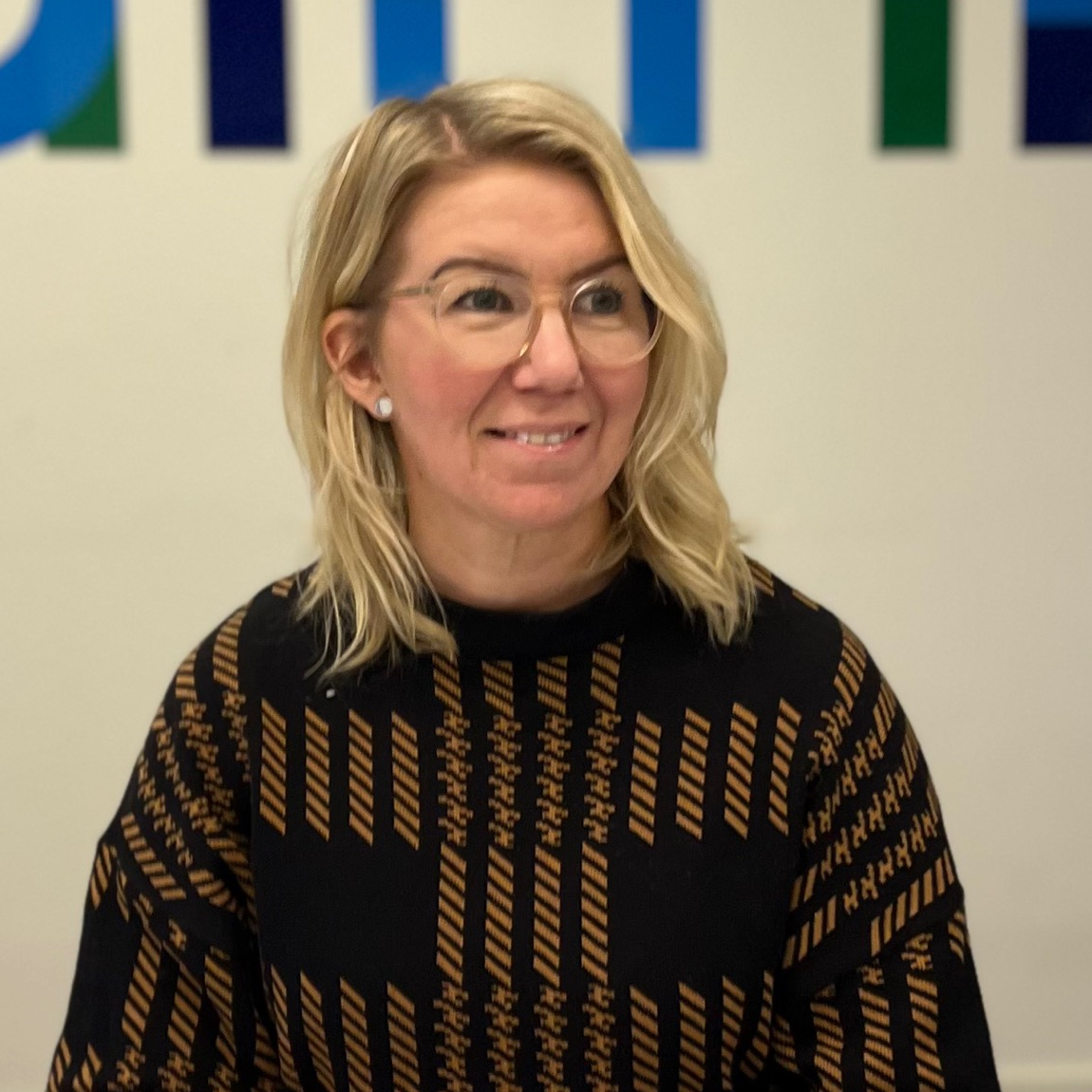 Porträtt av en kvinna med blont axellångt hår och glasögon. Hon bär en svart tröja med brunt mönster och sitter framför en ljus vägg med delar av en färgglad logotyp av Almi i bakgrunden. Hon ler lätt och tittar snett åt sidan.