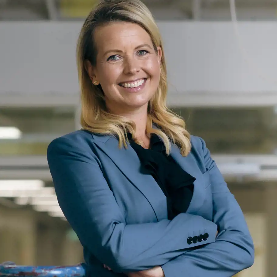 En leende kvinna i en blå kavaj och svart blus står med armarna i kors i en ljus kontorsmiljö. Hon ser självsäker ut och tittar rakt in i kameran.