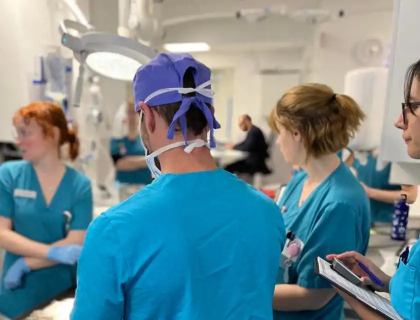 Flera sjukvårdspersonal i blå arbetskläder samlas runt en patient i en operations- eller vårdsal. En person i förgrunden bär kirurgmössa och munskydd, medan en annan antecknar på en skrivplatta. Miljön är klinisk med medicinsk utrustning synlig i bakgrunden.