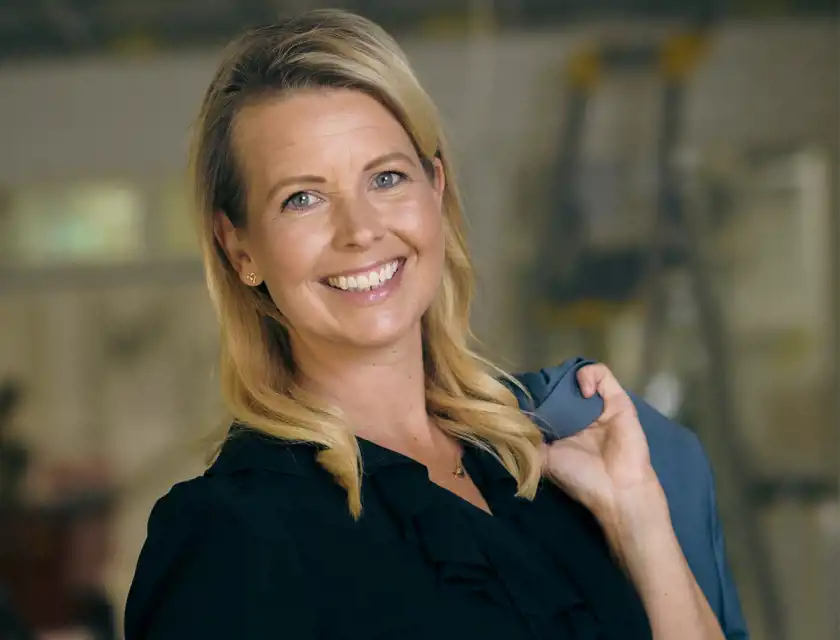 Leende kvinna iklädd svart blus, håller en blå kavaj över axeln. Bakgrunden är suddig men antyder en inomhusmiljö, möjligen en arbetsplats eller kontor.