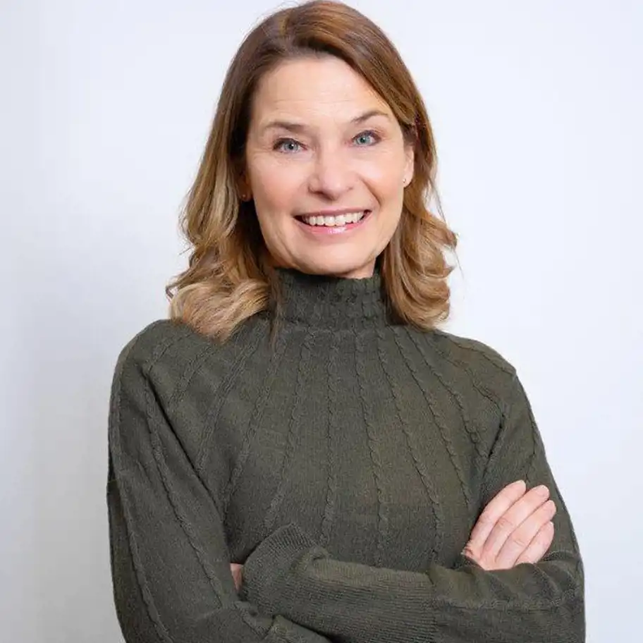 Kvinna med axellångt brunt hår och lockar i topparna står med armarna i kors och ler mot kameran. Hon bär en mörkgrön stickad tröja med hög krage. Bakgrunden är ljus och enkel.