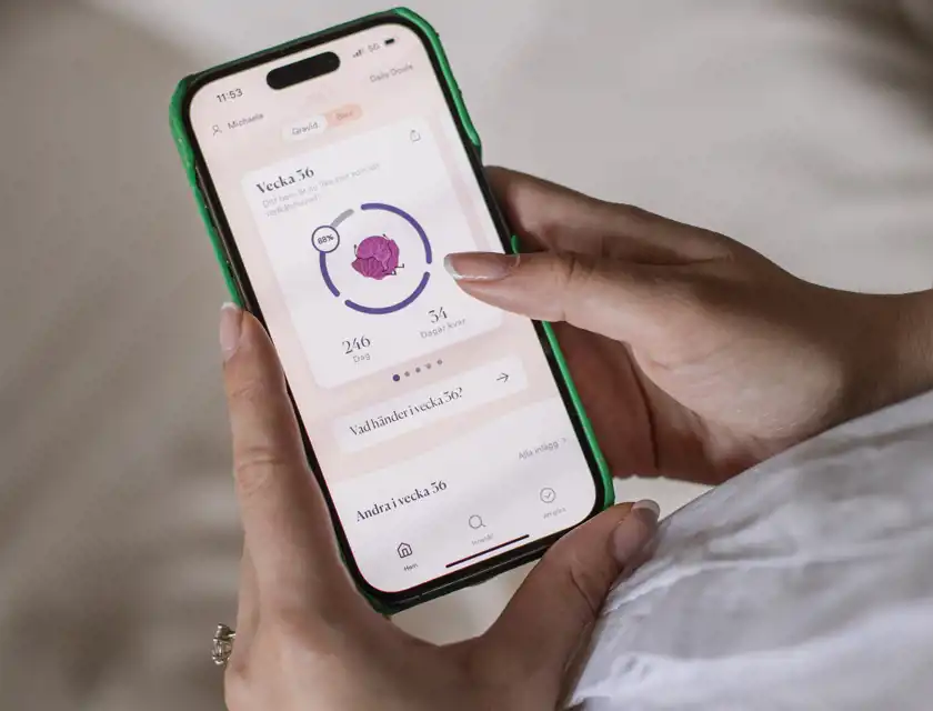 En person håller en smartphone med en graviditetsapp öppen. Skärmen visar att graviditeten är i vecka 36 och att barnet nu är lika stort som ett rödkålshuvud. Appen visar en cirkeldiagramindikator på 88 %, samt information om att 246 dagar har gått och att 34 dagar återstår. Personen har välmanikyrerade naglar och en förlovningsring på vänster hand. Bakgrunden är suddig och ser ut att vara en ljus säng eller soffa, vilket skapar en avslappnad och mysig känsla.