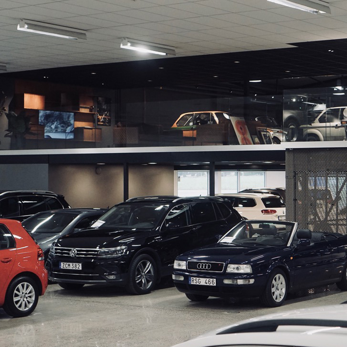 En modern bilhall med flera bilar i olika modeller och färger, inklusive en svart Volkswagen SUV och en mörkblå Audi cabriolet i förgrunden. Lokalen har en stilren inredning med dämpad belysning och en övervåning där klassiska bilar och bilder är utställda bakom glas.