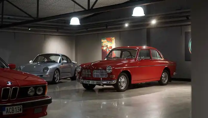 En stilfull bilhall med en klassisk röd Volvo Amazon i fokus, omgiven av andra veteranbilar, inklusive en silverfärgad Porsche 911. Lokalen har en modern belysning som framhäver bilarnas eleganta linjer, och en färgglad tavla hänger på väggen i bakgrunden.