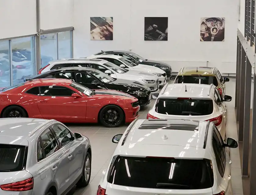 En modern bilhall med en rad bilar i olika modeller och färger, inklusive en iögonfallande röd Chevrolet Camaro. Stora fönster släpper in naturligt ljus, och på väggarna hänger stilrena bilder av bilinteriörer. Lokalen har en ren och välorganiserad layout, med bilarna parkerade i raka rader.