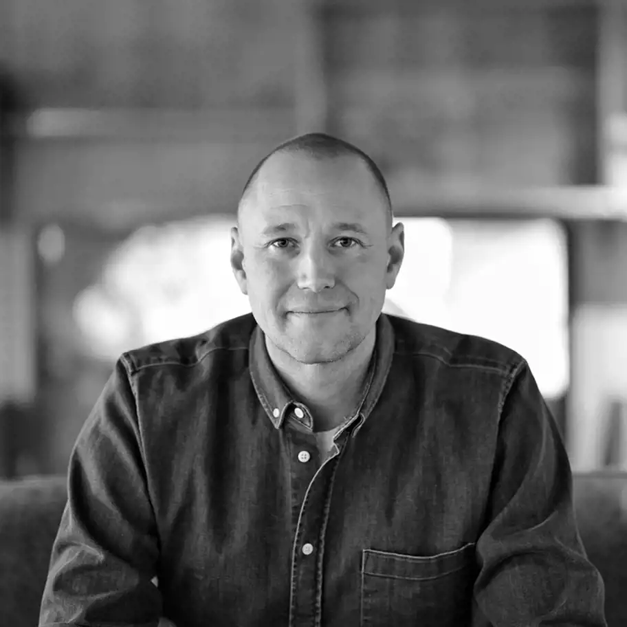 Porträtt i svartvitt av en man med rakat huvud och ett vänligt uttryck. Han bär en mörk jeansskjorta och sitter avslappnat i en inomhusmiljö med en mjukt oskarp bakgrund som ger en varm och inbjudande känsla.