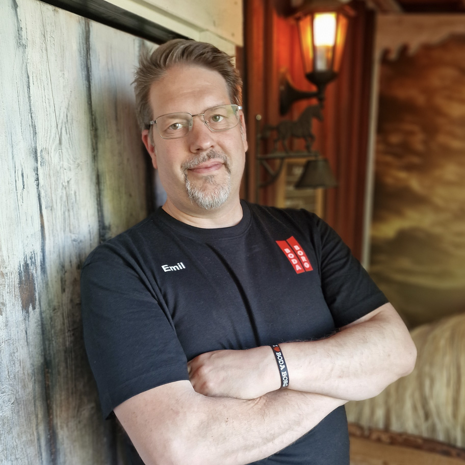 Man med glasögon och gråsprängt skägg lutar sig avslappnat mot en trävägg med armarna i kors. Han bär en svart t-shirt med namnet "Emil" tryckt på bröstet och ett armband med texten "BODA BORG". Bakgrunden har en rustik känsla med en gammaldags lykta, en hästdekoration och en tavla med en dramatisk himmel.