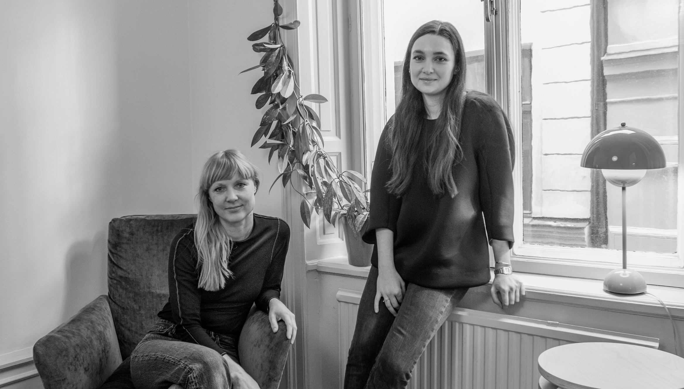En svartvit bild där två kvinnor sitter i ett ljust rum. Den ena sitter i en fåtölj och den andra sitter på en fönsterbräda. En växt hänger vid fönstret och en bordslampa står bredvid dem.
