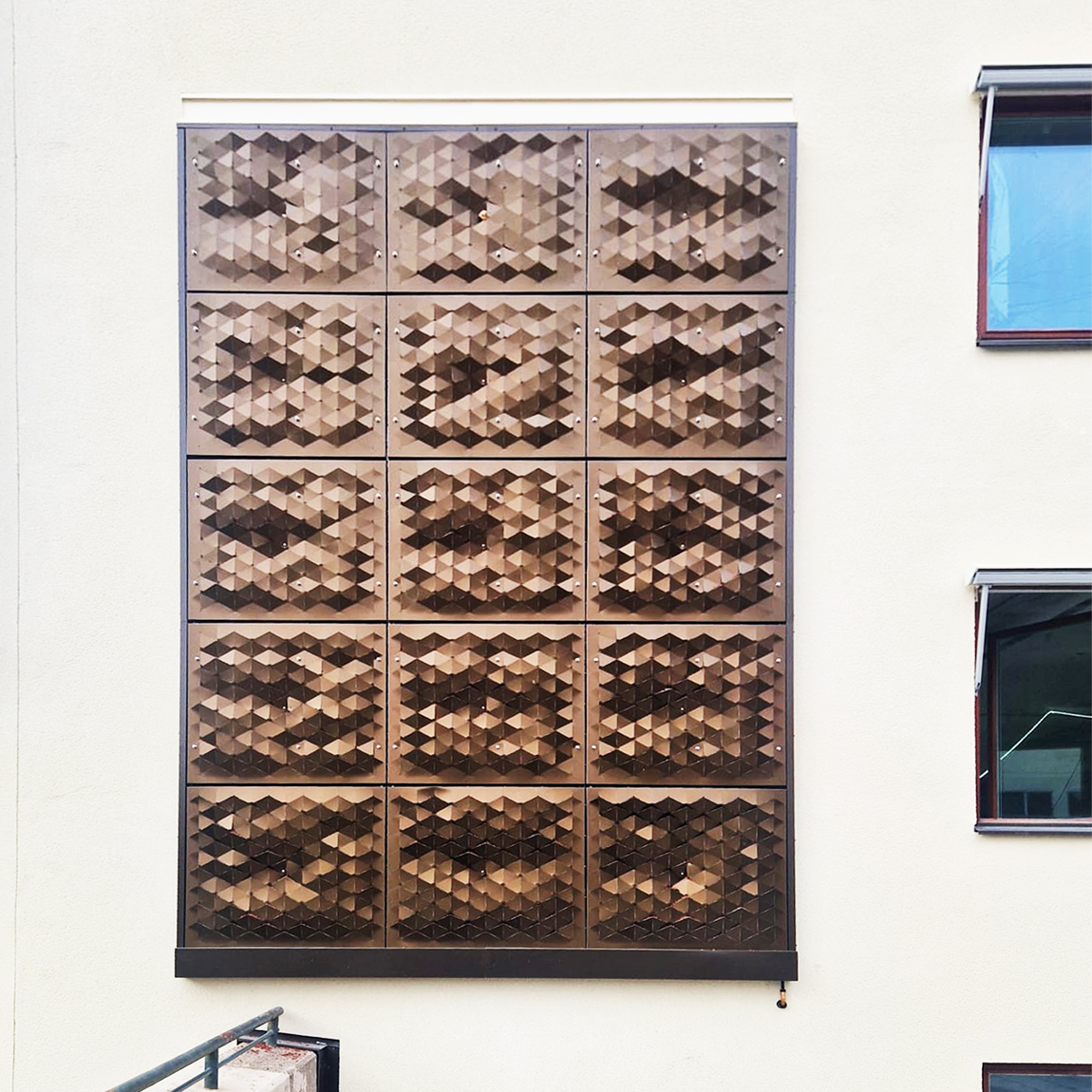 Butongs fasadinstallation med fasettformade metallpaneler monterade i ett rutmönster på en ljus yttervägg.