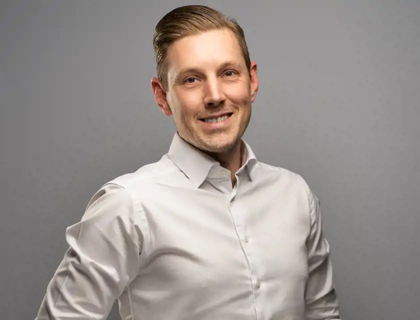 En man med bakåtkammat hår och en vit skjorta ler mot kameran mot en grå bakgrund.