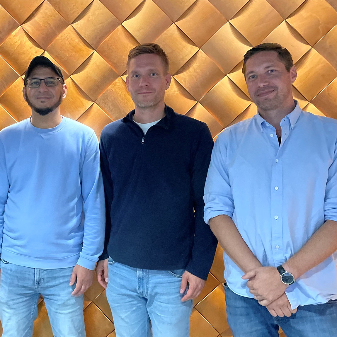 Tre män från företaget CareIfAI står framför en vägg med ett mönster av ljusa, vågformade träpaneler. De ler mot kameran och står axel vid axel i en avslappnad miljö.