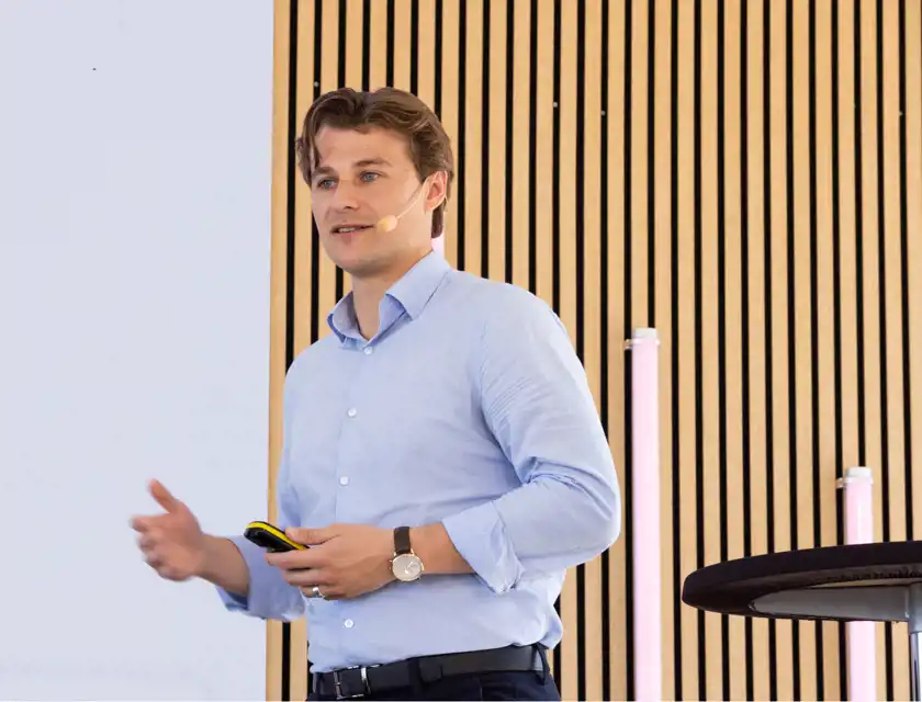 En person i en ljusblå skjorta håller en presentation på en scen. Personen har en mikrofon fäst vid kinden och håller en presentationskontroll i handen. Bakgrunden består av en vägg med vertikala träpaneler och några upplysta rör i rosa nyanser.